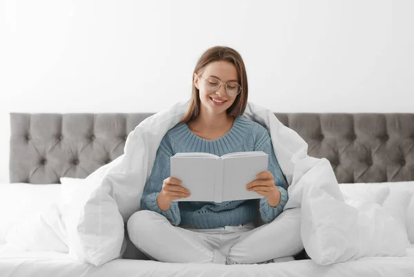 Ung kvinna i varm tröja läser bok på sängen hemma — Stockfoto