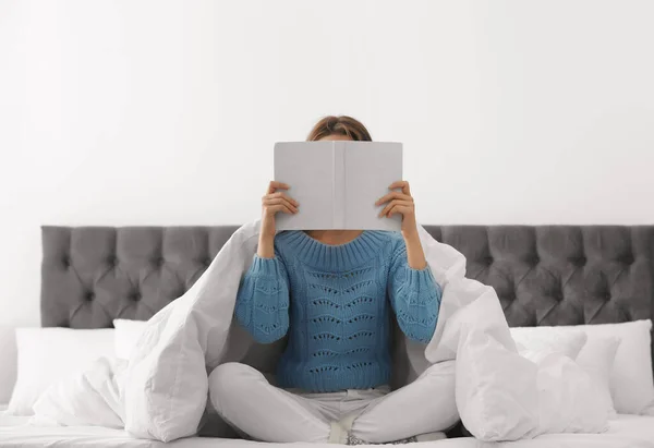 Młoda kobieta w ciepłym swetrze czyta książkę na łóżku w domu — Zdjęcie stockowe