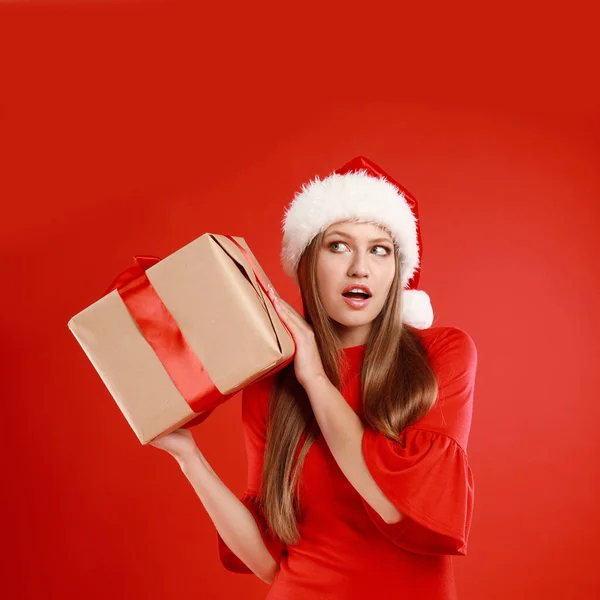 Noel Baba şapkalı, kırmızı arka planda Noel hediyesi olan duygusal genç bir kadın. — Stok fotoğraf