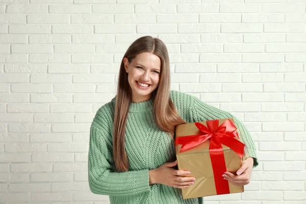 Ευτυχισμένη νεαρή γυναίκα με χριστουγεννιάτικο δώρο κοντά στον τοίχο από λευκό τούβλο — Φωτογραφία Αρχείου