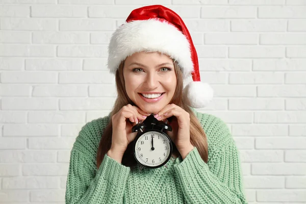 Joyeux jeune femme dans le chapeau de Père Noël avec réveil près du mur de briques blanches. Temps de Noël — Photo