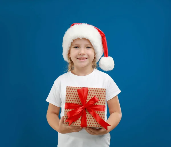 Bonito criança usando chapéu de Papai Noel com presente de Natal no fundo azul — Fotografia de Stock