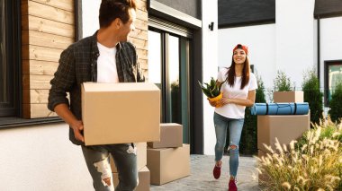Güneşli bir günde yeni evlerinin yakınında kutuları ve ev eşyaları taşıyan mutlu bir çift.