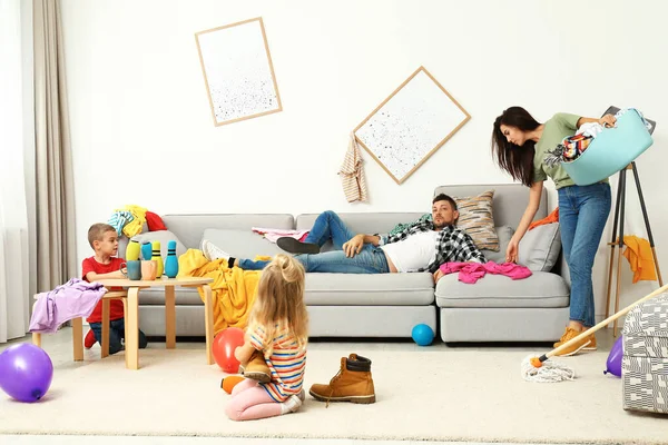 Exhausted mother trying to clean up mess made by children and lazy father in room — Stock Photo, Image