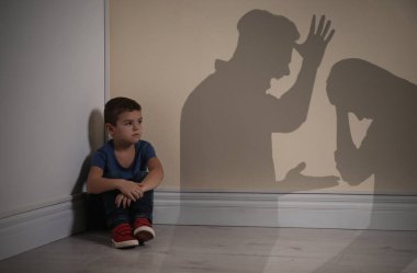 Sarı duvarın yanında oturan küçük çocuk ve tartışan ebeveynlerin siluetleri. 