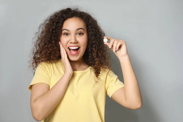 Emotional African-American woman with vitamin pill on light grey background