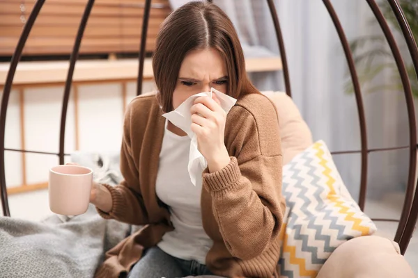Sick young woman with cup of hot drink sneezing at home. Influenza virus