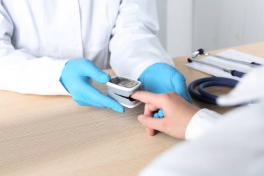 Doktor, modern parmak ucu nabız oksimetresi olan hastayı ahşap masada muayene ediyor.