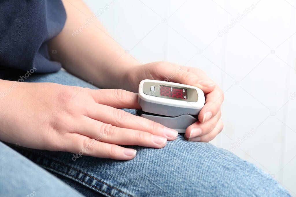 Woman using fingertip pulse oximeter on light background, closeup