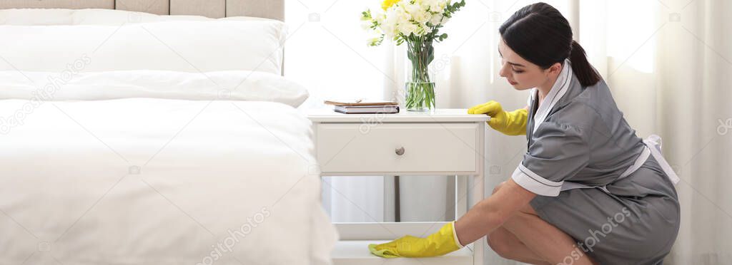 Young chambermaid wiping dust from nightstand in bedroom. Banner design