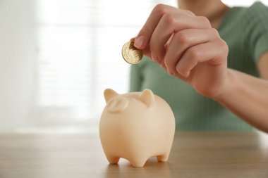 Woman putting money into piggy bank at wooden table indoors, closeup. Space for text clipart