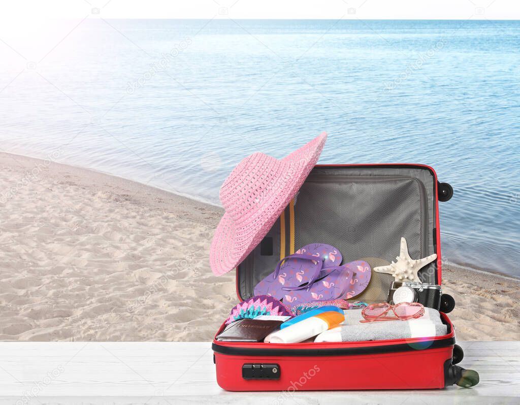 Suitcase with different beach objects on wooden table near sea. Space for text