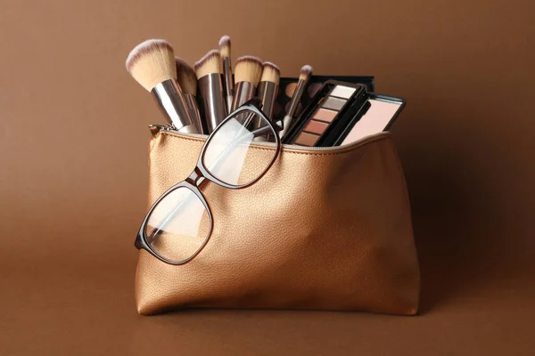 Cosmetic bag with makeup products and eyeglasses on brown background