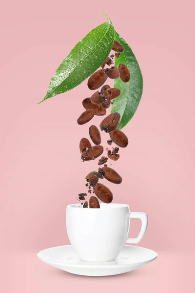 Cocoa beans and leaves falling into cup on pink background