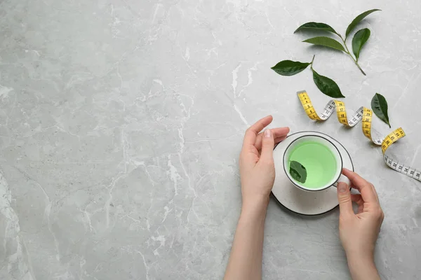 Woman with cup of herbal diet tea and measuring tape at light table, top view. Space for text