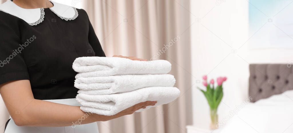 Young maid holding stack of fresh towels in hotel room, closeup view with space for text. Banner design