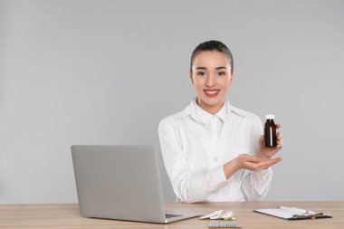 Açık gri arka plana karşı masada şurup ve dizüstü bilgisayarlı profesyonel bir eczacı.