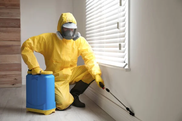 Pest control worker in protective suit spraying pesticide near window indoors