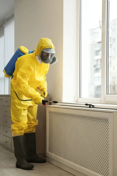 Pest control worker in protective suit spraying pesticide near window indoors