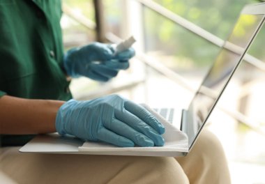 Lastik eldivenli bir kadın laptopunu ıslak mendille temizliyor.