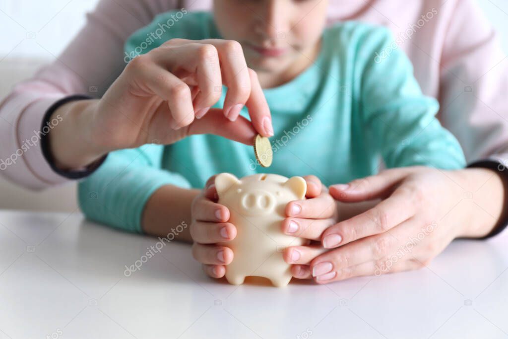 Mother and son putting coin into piggy bank at table indoors, closeup
