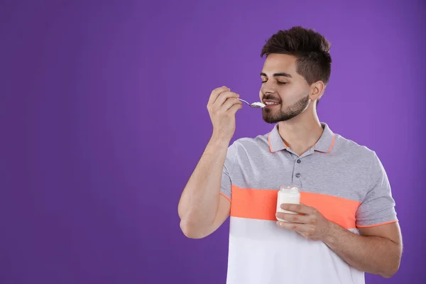 Happy young man eating tasty yogurt on purple background. Space for text