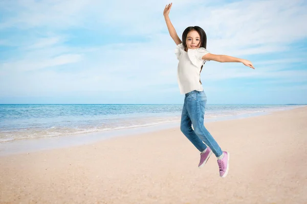 Cute school girl jumping on beach near sea, space for text. Summer holidays