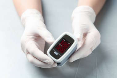 Parmak ucu nabız oksimetresi tutan eldivenli doktor açık gri masada, yakın plan.