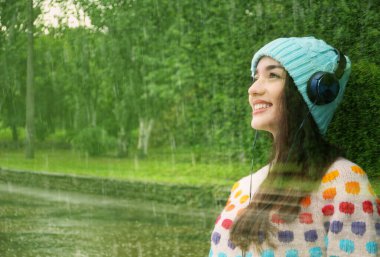Yeşil parkta sağanak yağmur ve müzik dinleyen kulaklıklı güzel bir kadın.