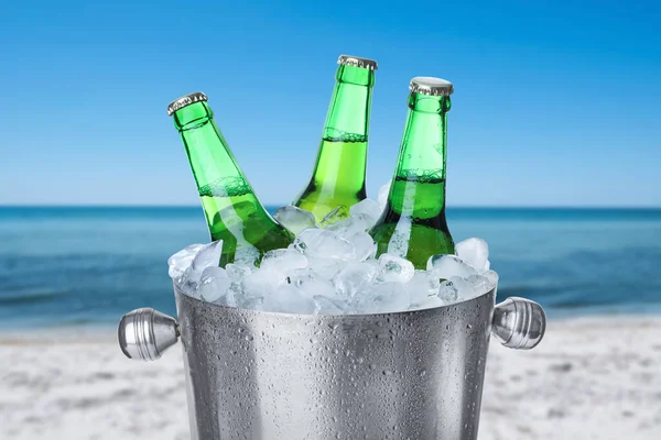 Bottles of beer with ice cubes in metal bucket against blurred sea and sandy beach