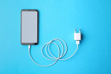 Açık mavi arka planda akıllı telefon ve USB şarj aleti, düz konum. Modern teknoloji