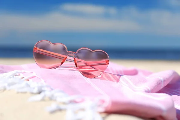 Stylish heart shaped sunglasses and blanket on sand near sea, closeup. Beach accessories for summer vacation
