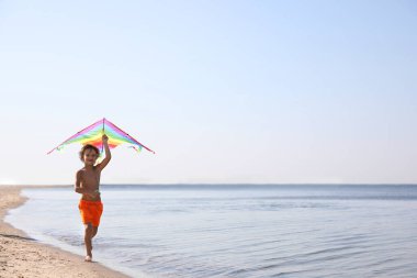 Güneşli bir günde kumsalda koşan uçurtmalı şirin bir çocuk.