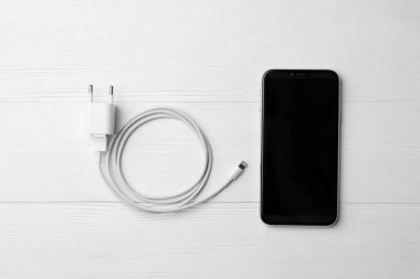 Akıllı telefon ve USB şarj aleti beyaz ahşap masada, düz yerde. Modern teknoloji
