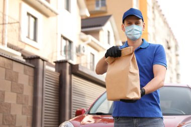 Koruyucu maskeli ve eldivenli bir kurye dışarıda arabanın yanında bekliyor. Coronavirus karantinası sırasında yiyecek teslimatı