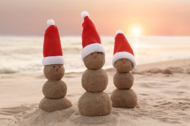 Gün batımında deniz kenarında kumdan yapılmış Noel Baba şapkalı kardan adamlar. Noel tatili