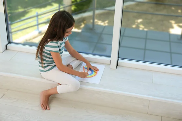 Little girl drawing rainbow near window indoors. Stay at home concept
