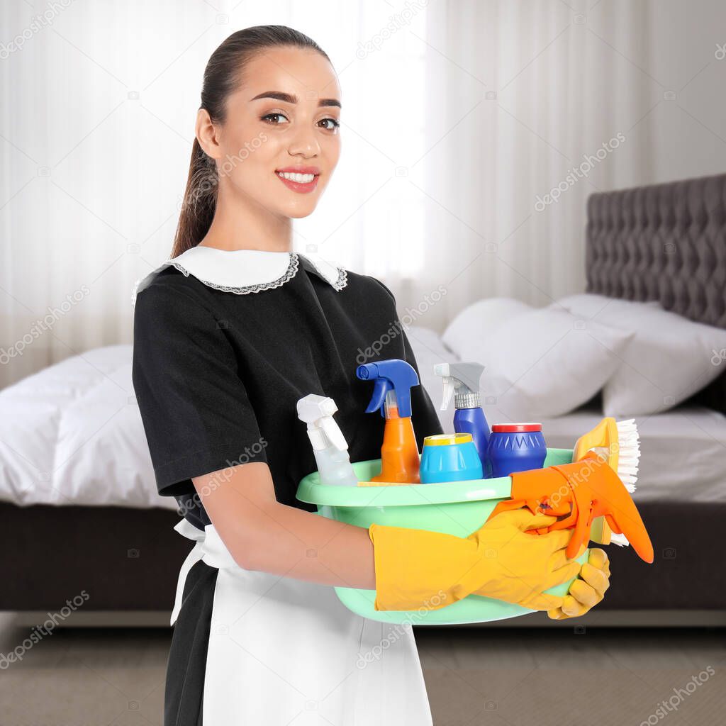 Beautiful chambermaid with cleaning supplies near bed in hotel room
