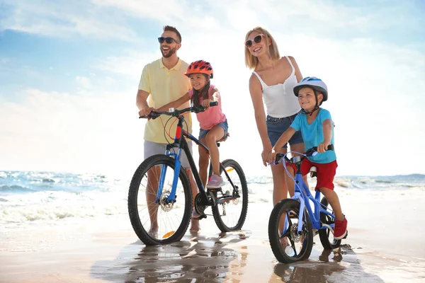 Happy parents teaching children to ride bicycles on sandy beach near sea