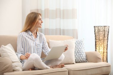 Evdeki kanepede dizüstü bilgisayarlı güzel bir kadın oturuyor.