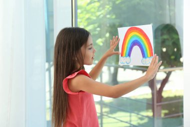 Little girl with picture of rainbow near window indoors.  Stay at home concept clipart
