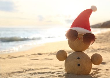 Noel Baba şapkası ve güneş gözlükleriyle kumdan yapılmış bir kardan adam, deniz kenarında bir plajda, mesaj için yer var. Noel tatili