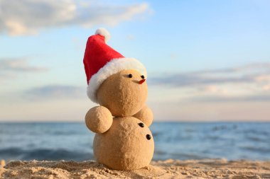 Kumsalda deniz kenarında Noel Baba şapkasıyla kumdan yapılmış bir kardan adam. Noel tatili