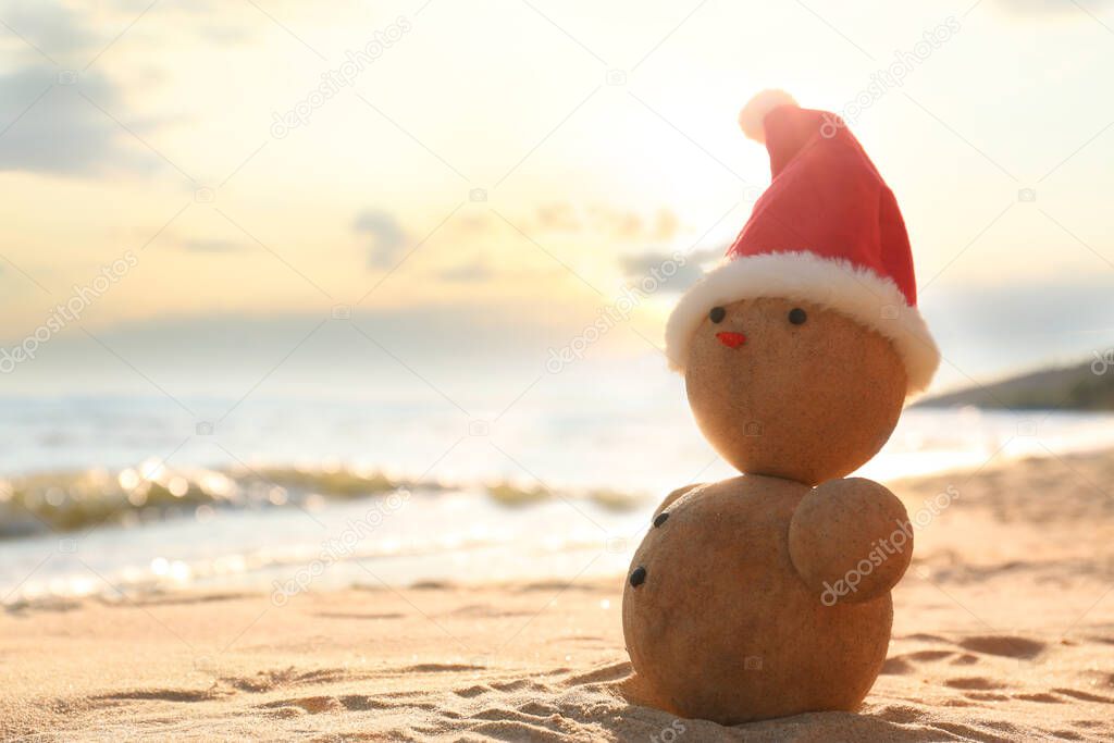 Snowman made of sand with Santa hat on beach near sea, space for text. Christmas vacation