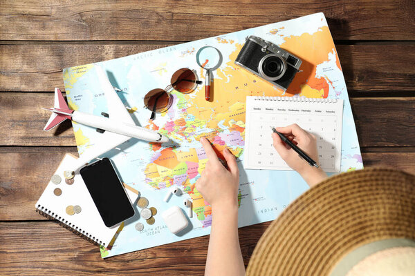Woman marking calendar at table with world map, top view. Travel during summer vacation