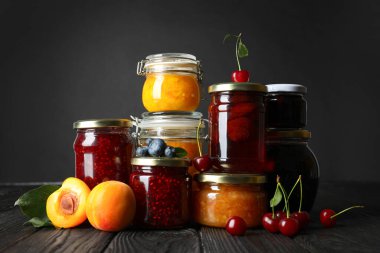 Jars with different jams and fresh fruits on black wooden table clipart