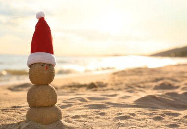 Gün batımında deniz kenarında kumdan yapılmış Noel Baba şapkalı bir kardan adam. Yazmak için yer var. Noel tatili