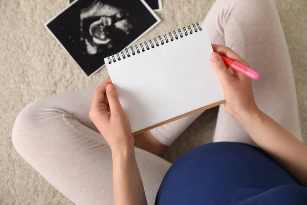 Pregnant woman with notebook and sonogram choosing name for baby on carpet, top view