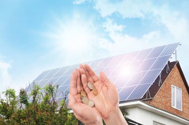 Man holding coins against house with installed solar panels. Renewable energy and money saving clipart