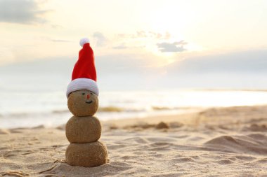 Gün batımında deniz kenarında kumdan yapılmış Noel Baba şapkalı bir kardan adam. Yazmak için yer var. Noel tatili